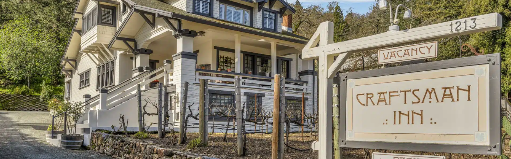 Front view of the Craftsman Inn in Calistoga, California, showcasing a charming white two-story house with dark trim, a wraparound porch, and a wooden sign reading “Craftsman Inn” near the driveway entrance.